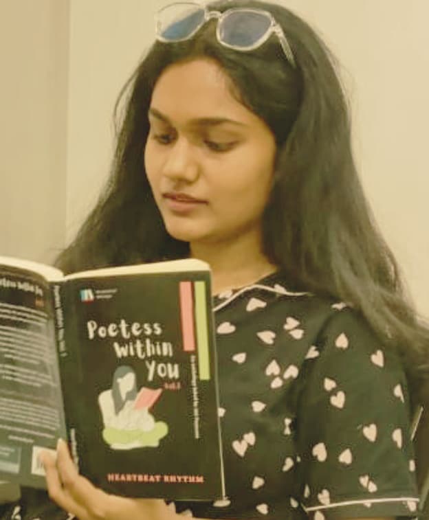 "Young woman reading the poetry book 'Poetess Within You' while seated indoors, wearing a heart-patterned top."
